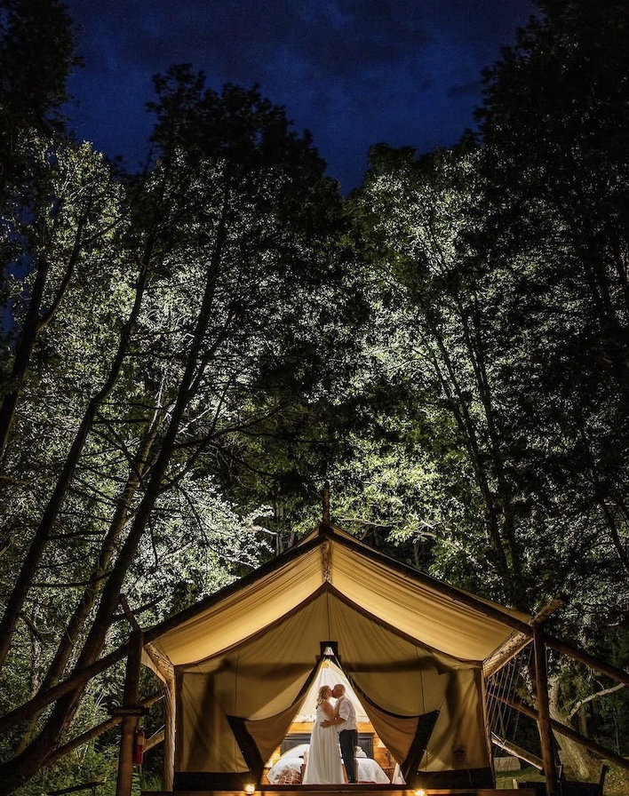 Kristina and Ron kissing in the doorway of a glowing tent beneath tall trees at night